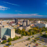 Downtown Topeka aerial skyline view with the Kansas State Capitol building in the center, and a blue sky with clouds in the background.